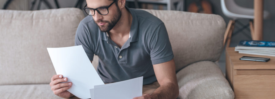 Time to look through bills. Handsome young man holding papers and looking at it while sitting on the couch at home